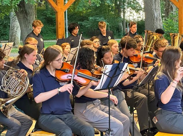 Student musicians performing in a mixed string and band ensemble during a live outdoor concert performance.