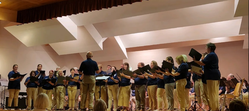 Adult choir performing on stage under the direction of a conductor during a live ensemble concert.