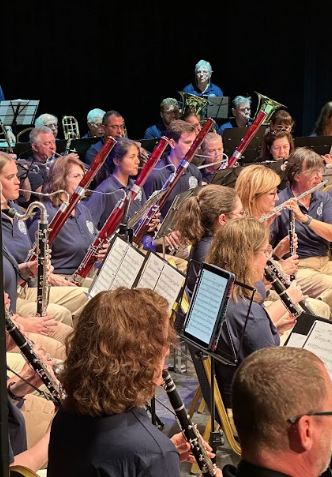 Adult community ensemble musicians performing with woodwind and brass sections during a live concert performance.