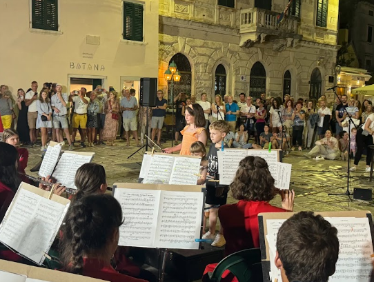 Student orchestra performing in a historic town square during an evening concert while local residents and visitors gather to watch.